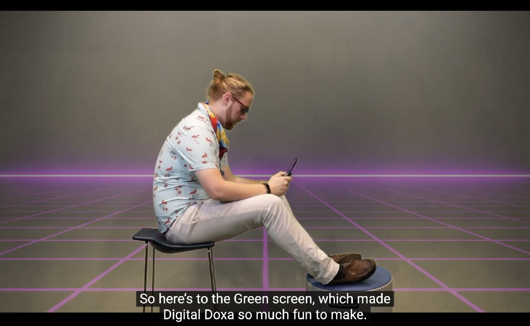 A white man sitting on a chair with his feet upon. The man is wearing a blue shirt with a flamingo pattern, red sunglasses, and a tie-dye bandana around his neck. He is staring down at the phone in his hands. The background is light grey and has a purple grid pattern on the ground.